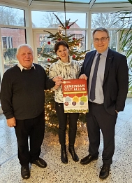 Helmut Sündermann, Ursula Seidler-Braun und Landrat Marco Prietz stehen vor einem geschmückten Weihnachtsbaum, halten ein Plakat in der Hand und schauen in die Kamera. © Landkreis Rotenburg (Wümme)