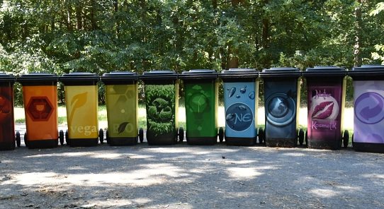Eine Reihen von bunt beklebten Mülltonnen steht auf einem betonierten Platz. Im Hintergrund sind Bäume und Rasen zu erkennen.