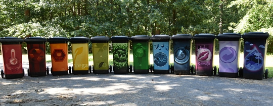 Eine Reihen von bunt beklebten Mülltonnen steht auf einem betonierten Platz. Im Hintergrund sind Bäume und Rasen zu erkennen. © Landkreis Rotenburg (Wümme)