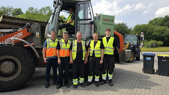 Fünf Männer in Arbeitskleidung stehen vor einem Bagger, in dem ein weitere Mann sitzt. Neben dem Bagger stehen eine blaue und eine gelbe Tonne.